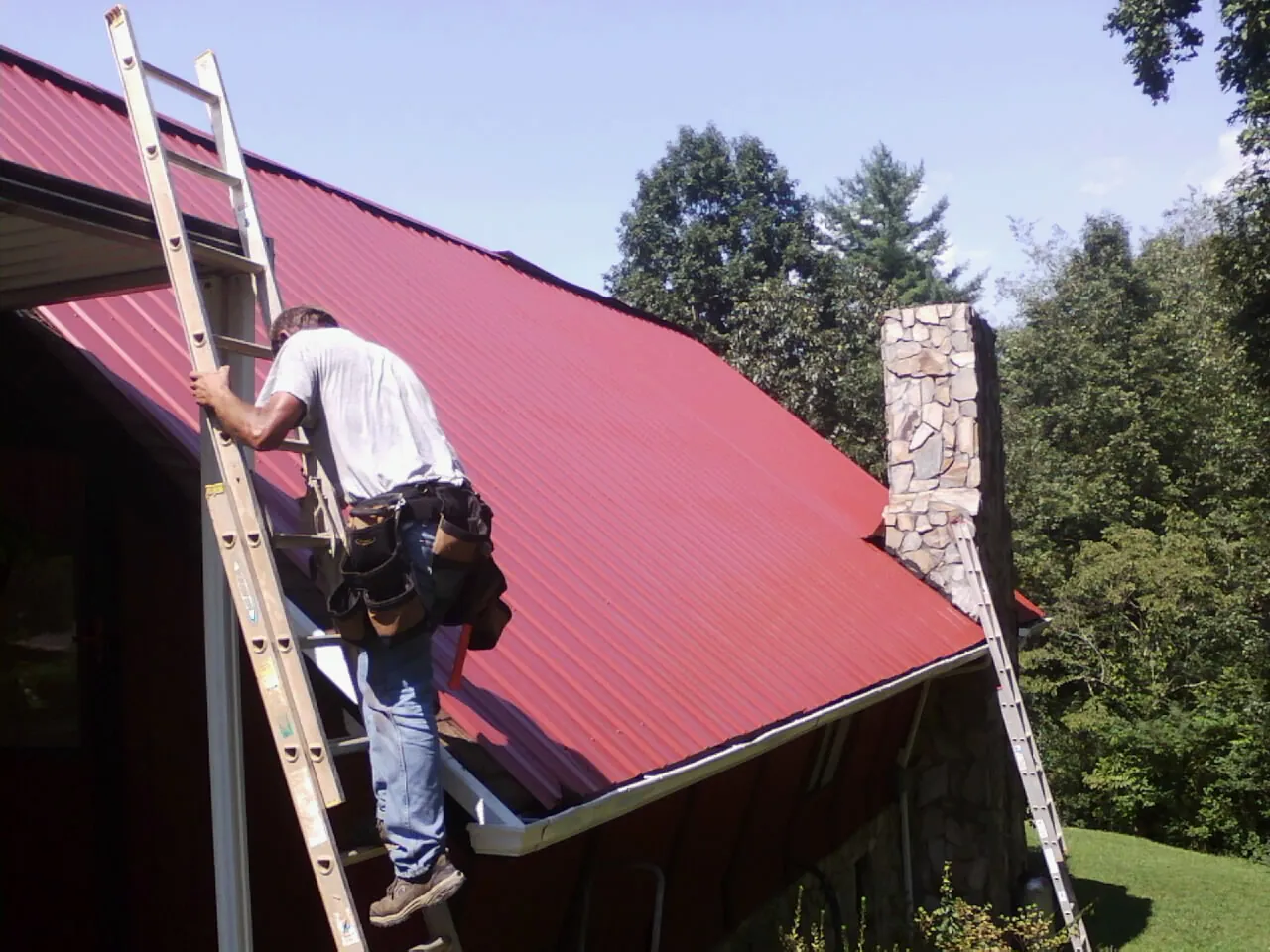 Roofer on a ladder beside a red metal roof and stone chimney — Soffit & Fascia Repair by Roofing Brothers in Arlington